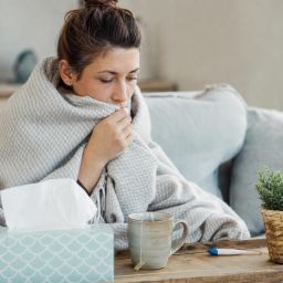 Woman wrapped in a blanket on the couch with tissues, tea and a thermometer