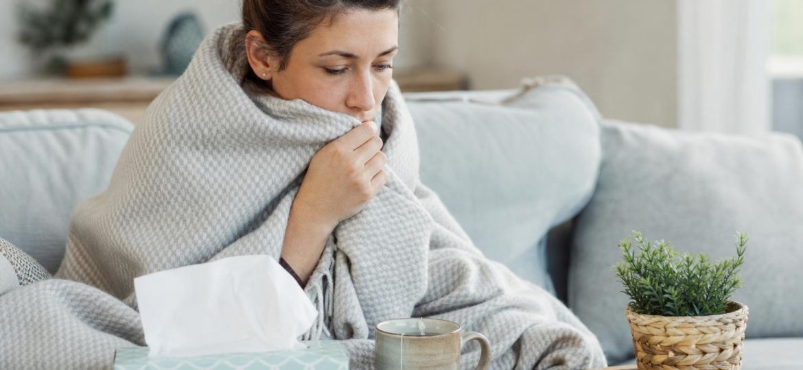 Woman wrapped in a blanket on the couch with tissues, tea and a thermometer