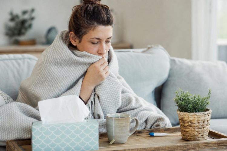 Woman wrapped in a blanket on the couch with tissues, tea and a thermometer