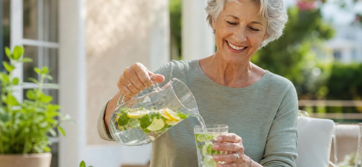 Older woman pouring citrus and mint water from a glass pitcher on a sunny patio