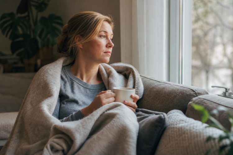 Woman resting on a sofa by a window with a blanket and a cup of tea