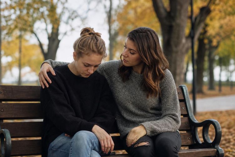 Friend comforting another with an arm around their shoulder on a park bench